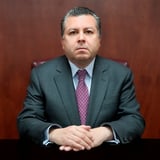Professional headshot of a middle-aged man in a dark suit and purple tie, seated against a burgundy background with hands folded