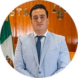 Professional headshot of a man in light blue blazer and dark tie, standing in front of wooden paneling with Mexican flag visible
