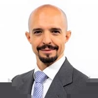 Professional headshot of a bald man with a beard wearing a dark suit and light blue checkered tie